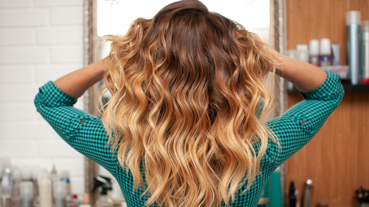 Von hinten fotografiert: Eine Frau mit Ombre-Strähnen in den Haaren wuschelt in ihre Haare einmal durch.