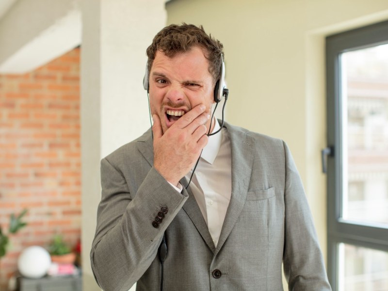 Ein Mitarbeiter in einem Callcenter mit Headset verzieht während eines Telefonats genervt das Gesicht.