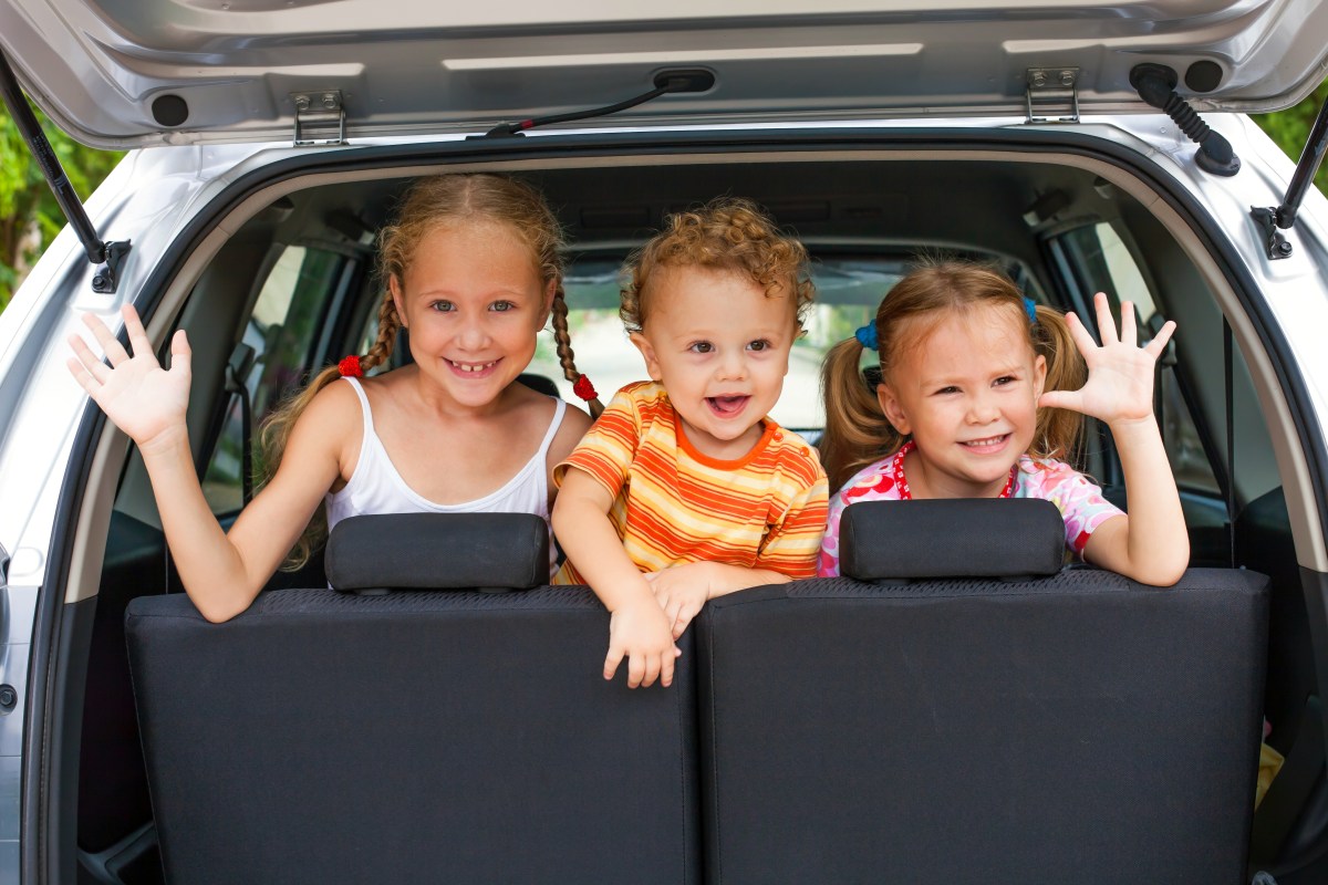 Drei kleine Kinder sitzen auf dem Rücksitz eines Autos und winken durch die geöffnete Kofferraum-Klappe.