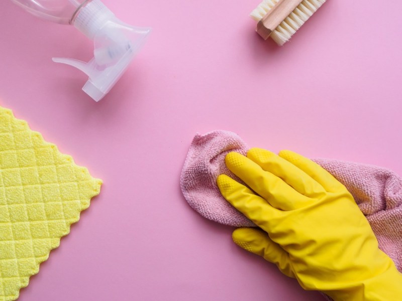 Utensilien zum Putzen und eine Hand mit einem gelben Gummihandschuh auf einem rosa Hintergrund