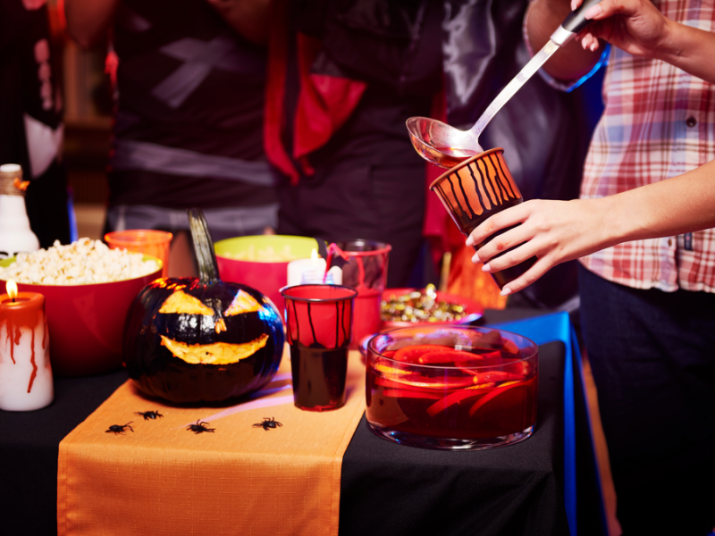 Ein Buffet auf einer Halloween-Party. Man sieht gruselige Deko und eine Frau gießt sich Bowle in einen Becher.