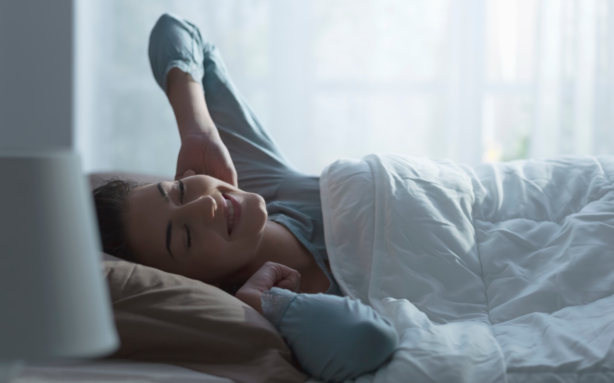 Eine junge Frau, die gerade aufgewacht ist, liegt im Bett und streckt sich gutgelaunt.