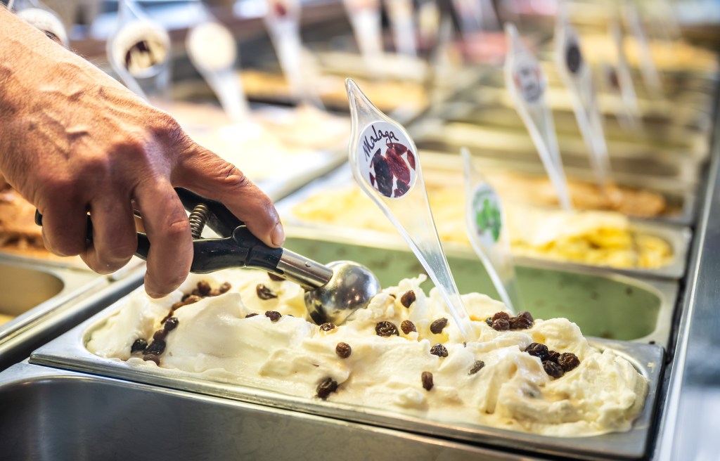 Eisverkäufer entnimmt eine Kugel Eis der Sorte „Malaga“.