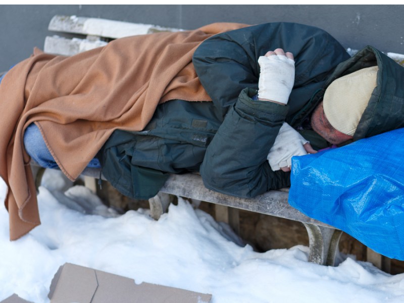 Ein obdachloser Mann schläft mit Decken bedeckt auf einer Bank im Winter.