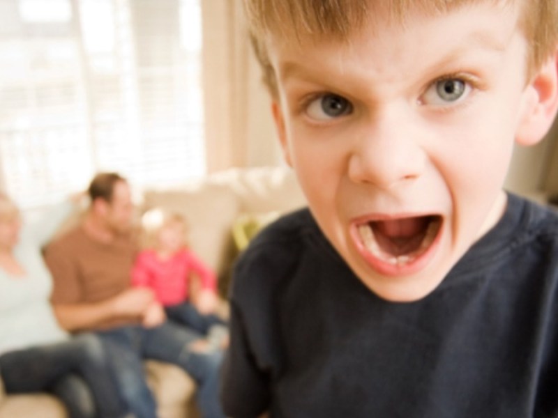 Kleiner Junge schreit in die Kamera, während hinter ihm seine Familie auf dem Sofa sitzt