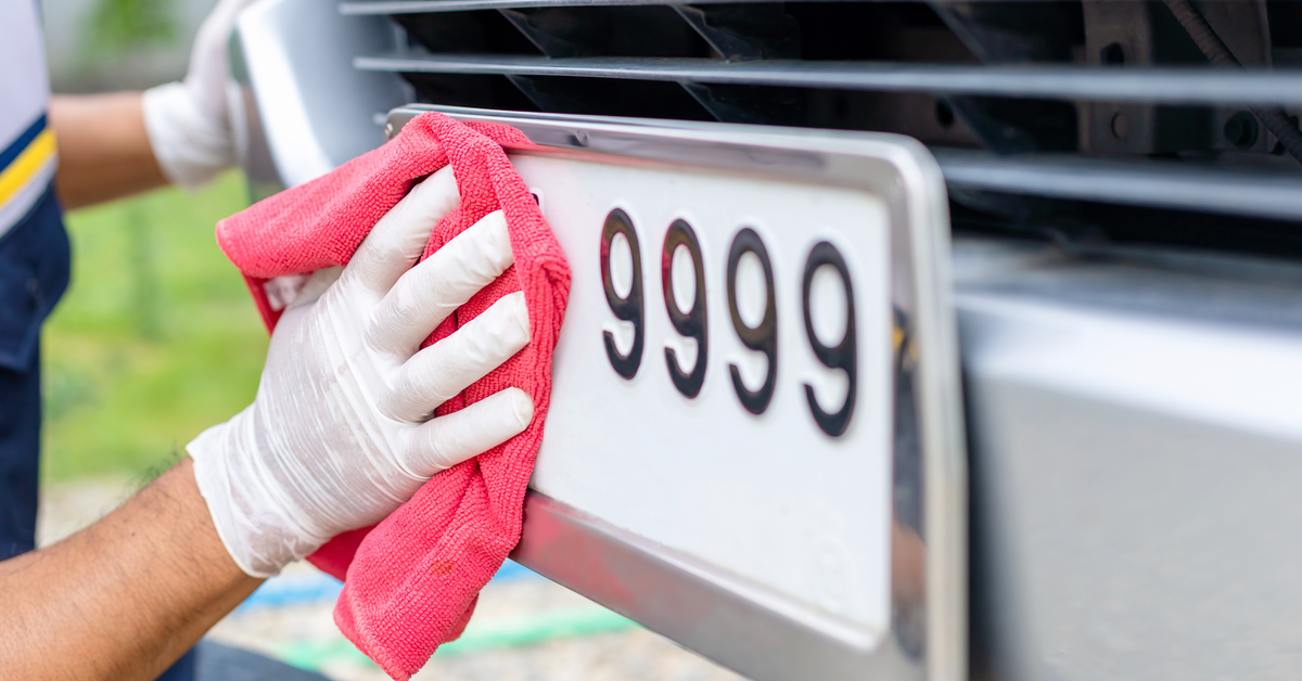 Eine Person säubert ein Nummerschild, das mit 9999 endet, mit einem roten Tuch. Dabei trägt die Person weiße Einmalhandschuhe.