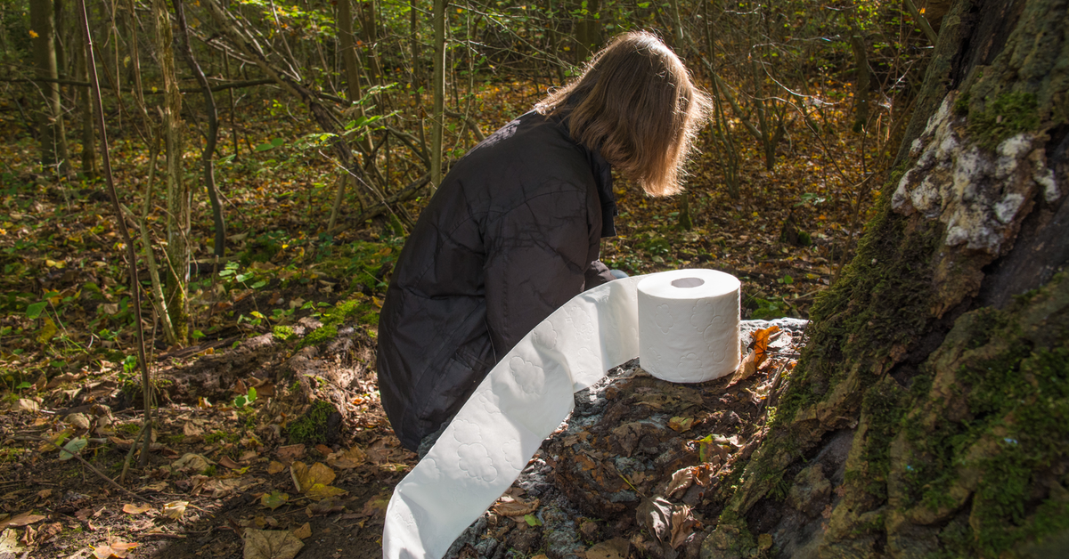 Eine Frau hockt neben einer Rolle Toilettenpapier in einem Wald.