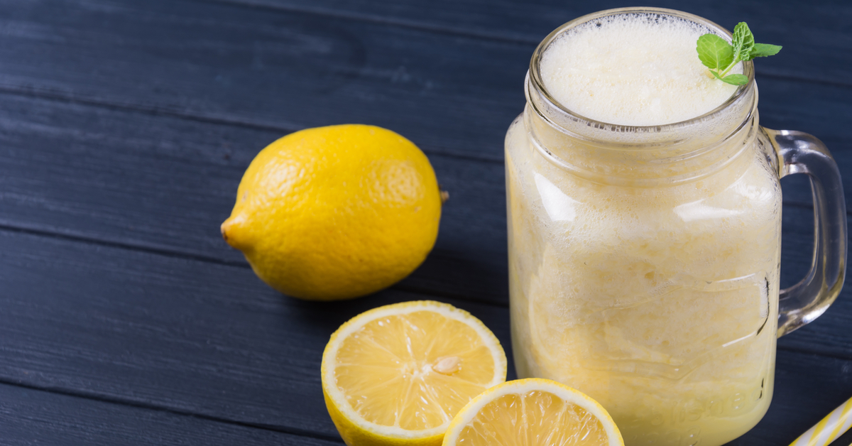 Ein Henkelglas mit Zitronenlimonade steht vor einem dunklen Hintergrund. Aufgeschnittene Zitronen liegen daneben.