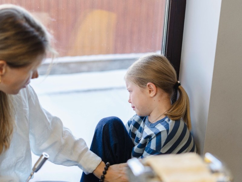 Mutter spricht auf Tochter ein, die traurig aus dem Fenster schaut