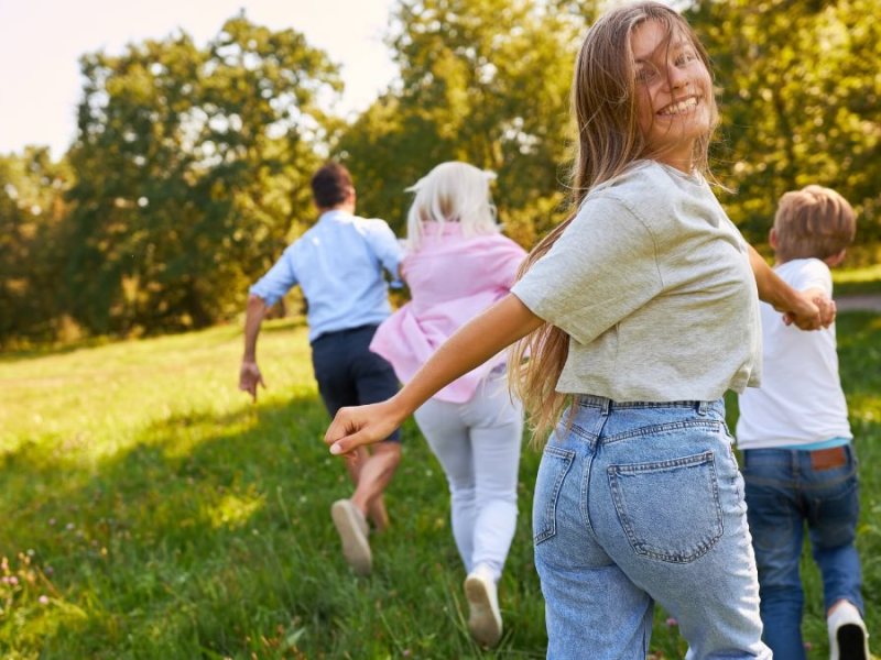 Eine Familie rennt frÃ¶hlich Ã¼ber eine Wiese. Das MÃ¤dchen hinten schaut glÃ¼cklich in die Kamera.