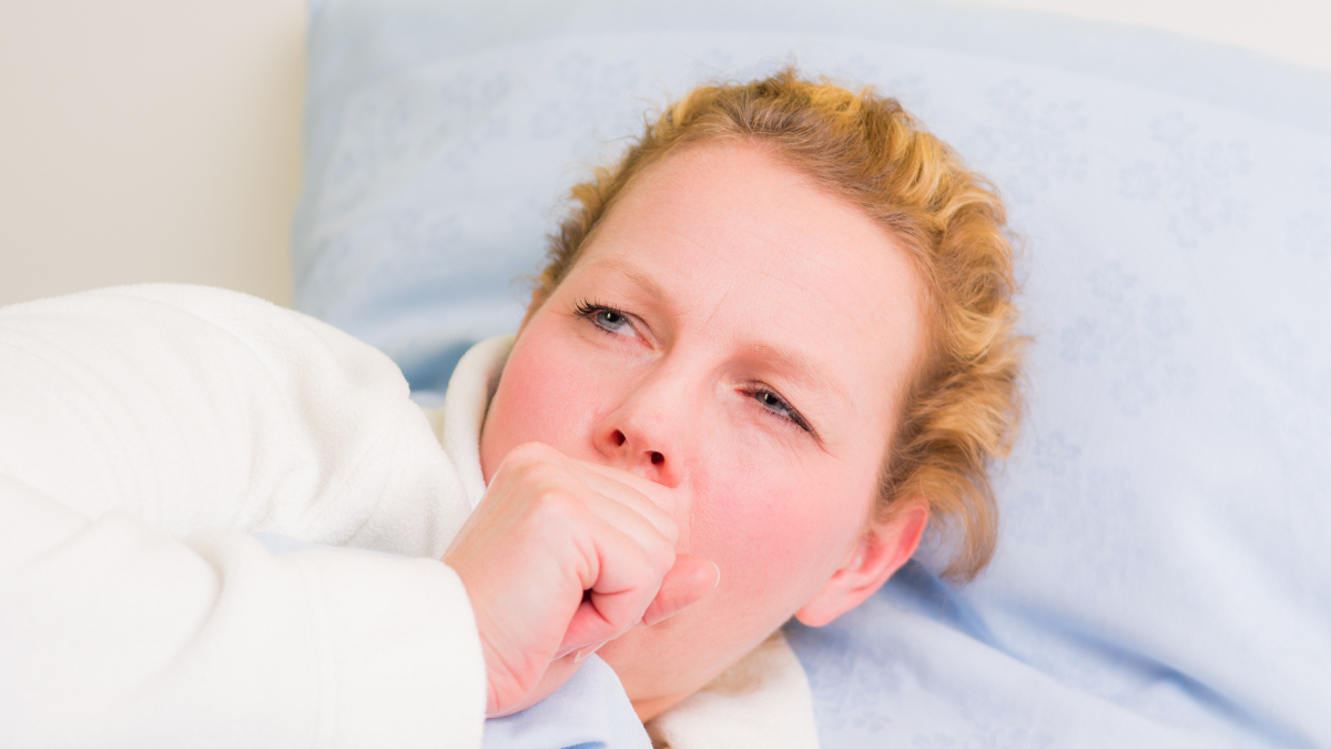 Eine blonde Frau im Bademantel liegt mit Husten im Bett.