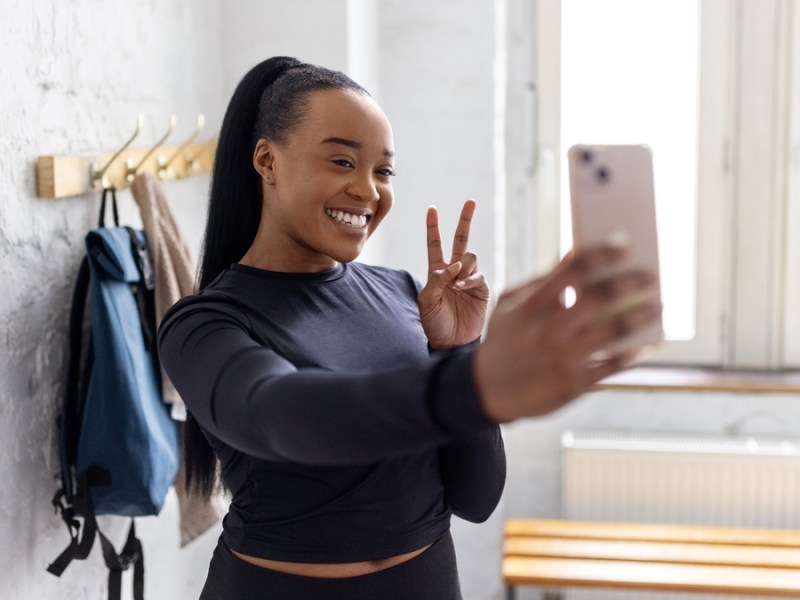 Eine Schwarze Frau mit einem Pferdeschwanz zeigt das Peace-Zeichen und macht ein Selfie.