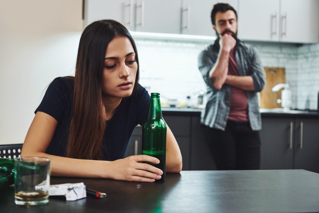 Eine betrunkene junge Frau sitzt vor einer Flasche Alkohol an einem Tisch und ihr besorgter Partner steht im Hintergrund.