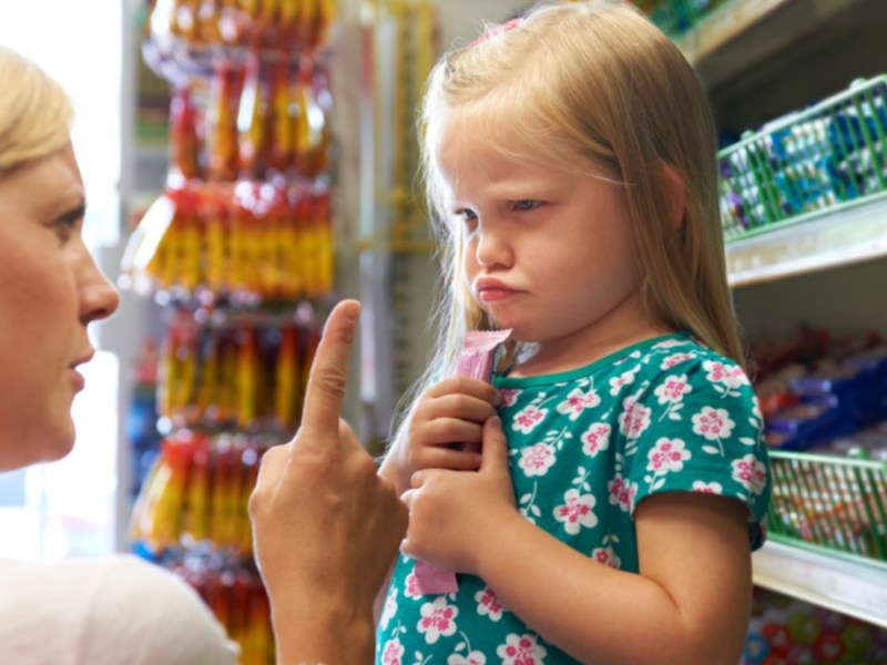 Eine Mutter schimpft mit erhobenem Zeigefinger mit einem Kind, das böse guckt und sich an eine Süßigkeit klammert.
