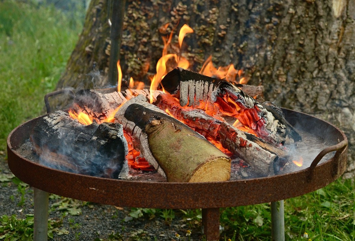 Eine Feuerschale, in der ein Feuer brennt, steht in einem Garten vor einem Baum.