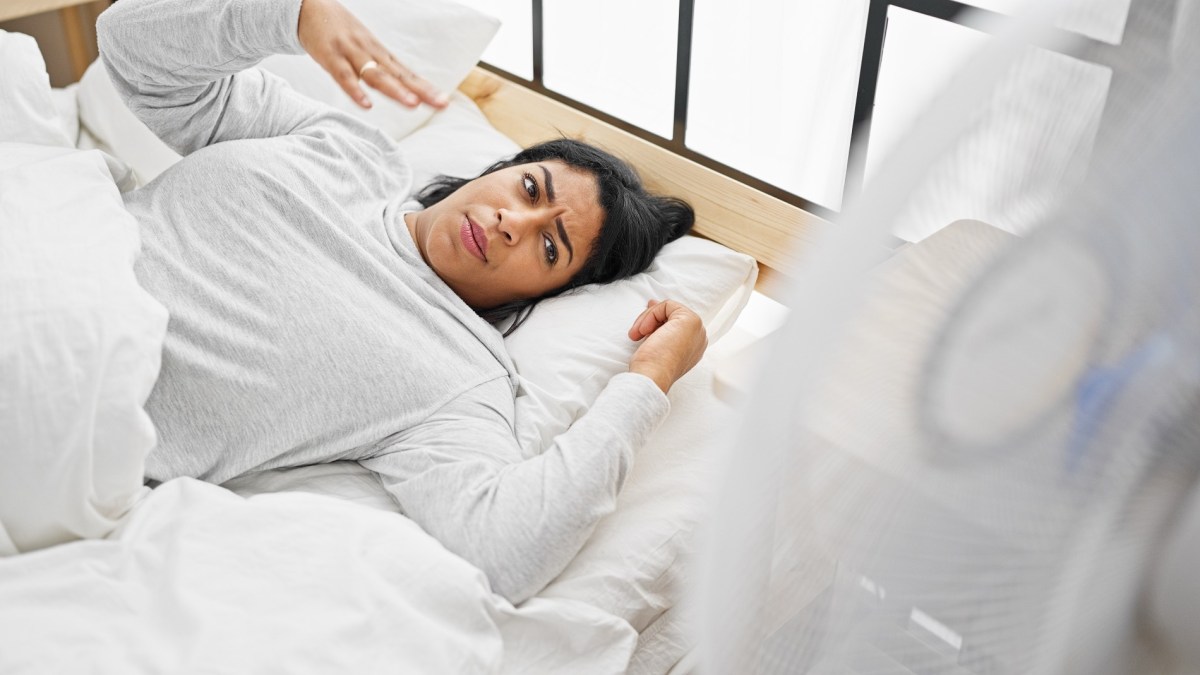 Eine Frau liegt in einem Bett mit Sommer-Bettwäsche und fächert sich kühle Luft zu.