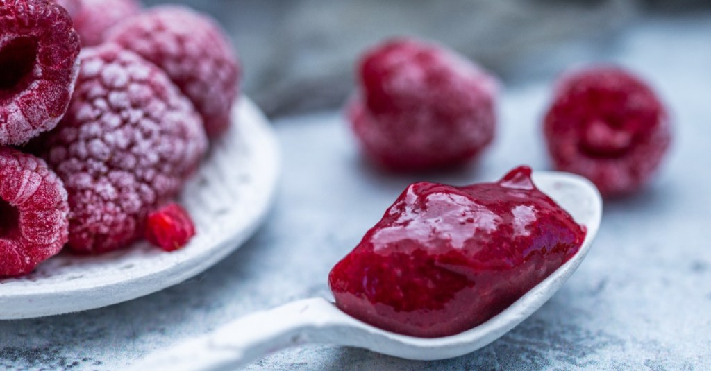 Ein Löffel mit Himbeermarmelade im Vordergrund, dahinter sind gefrorene Himbeeren zu sehen