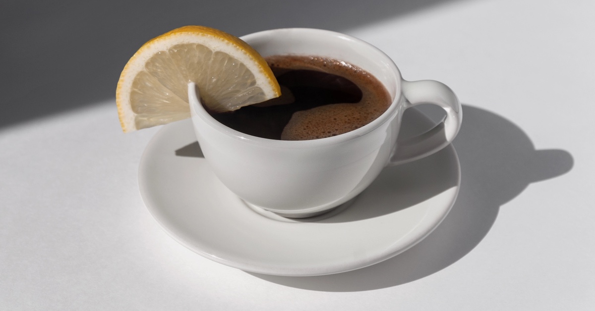 Tasse Kaffee steht samt Untertasse auf einem Tisch. Am Tassenrand befindet sich ein Stück Zitrone.