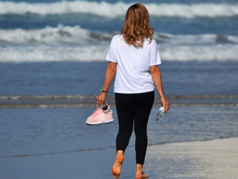 Eine Frau steht mit dem Rücken zur Kamera am Strand und blickt auf das Wasser.