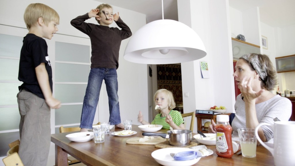 Eine Mutter sitzt am Tisch, während ihre zwei Söhne auf den Stühlen stehen und Grimassen machen.