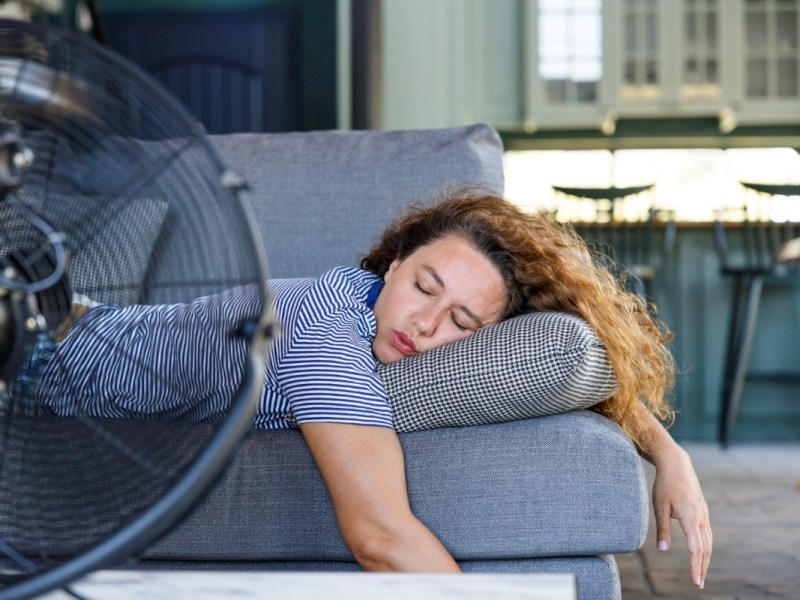 Ein Foto zeigt eine junge Frau beim Schlafen bei Hitze auf einer Couch, die sich mit einem Ventilator abkühlt.