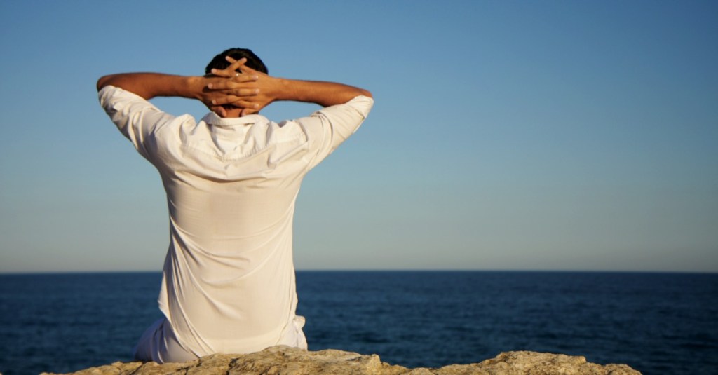 Mann sitzt alleine auf einem Felsen und schaut aufs Meer