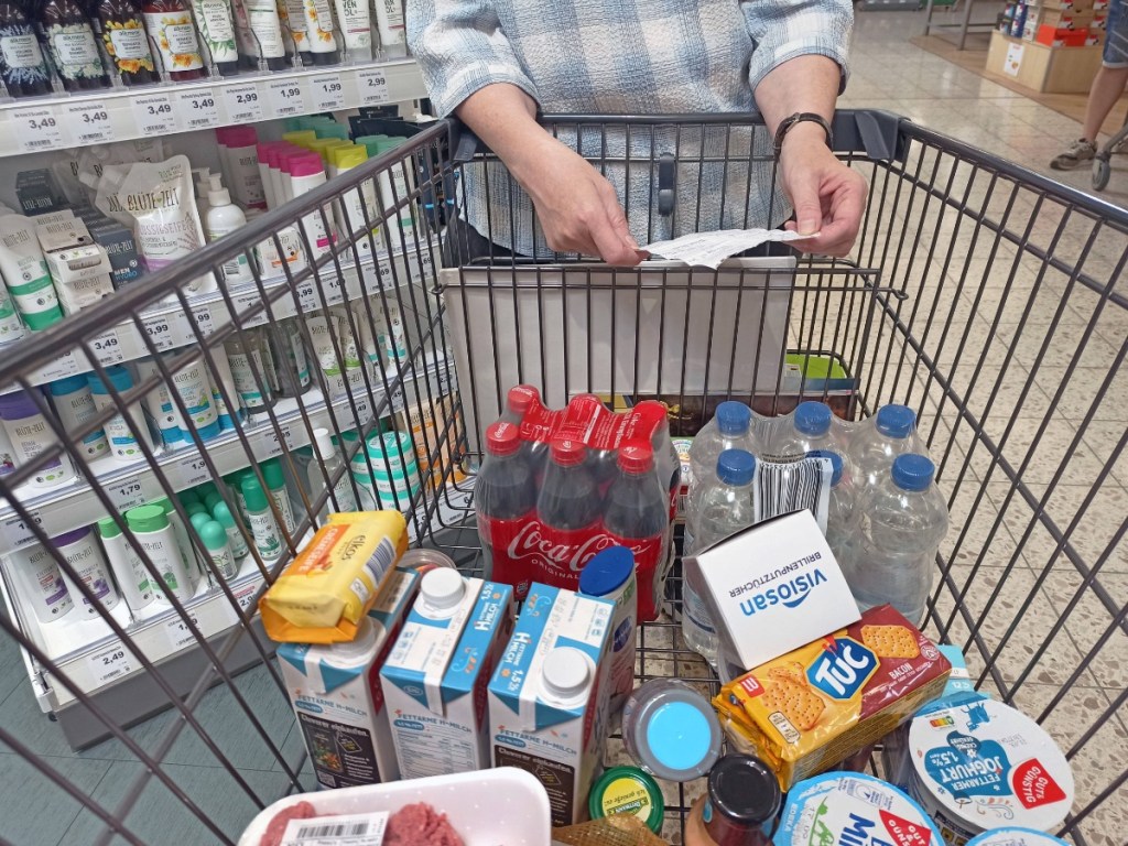 Frau mit Einkaufszettel stützt sich auf einen gut gefüllten Einkaufswagen.