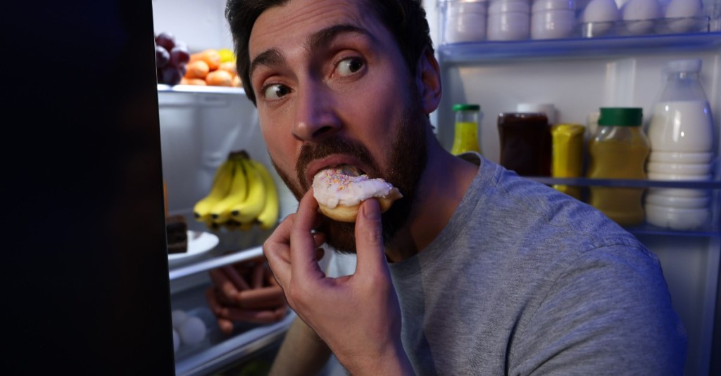 Mann isst heimlich einen Donut am offenen Kühlschrank