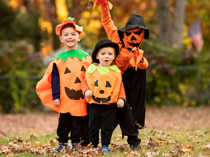 Drei Kinder mit Kürbiskostümen auf einer Wiese mit buntem Herbstlaub