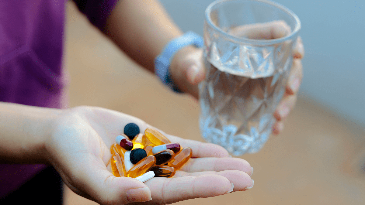 Eine Hand hält viele verschiedene Tabletten, die andere ein Glas Wasser.