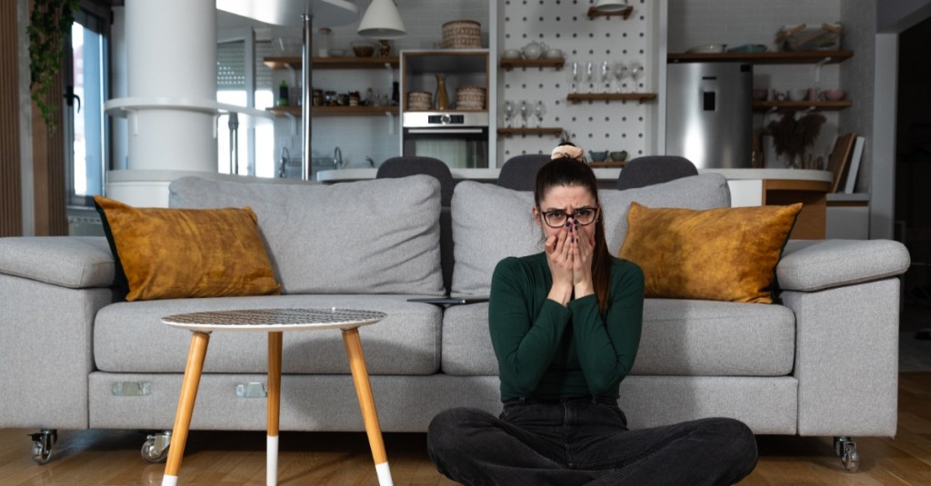 eine junge Frau sitzt vor ihrem Sofa und schlägt die Hände vors Gesicht