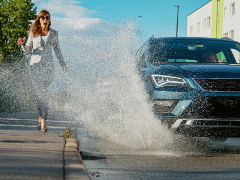 Auto fährt durch Pfütze und spritzt eine Fußgängerin nass