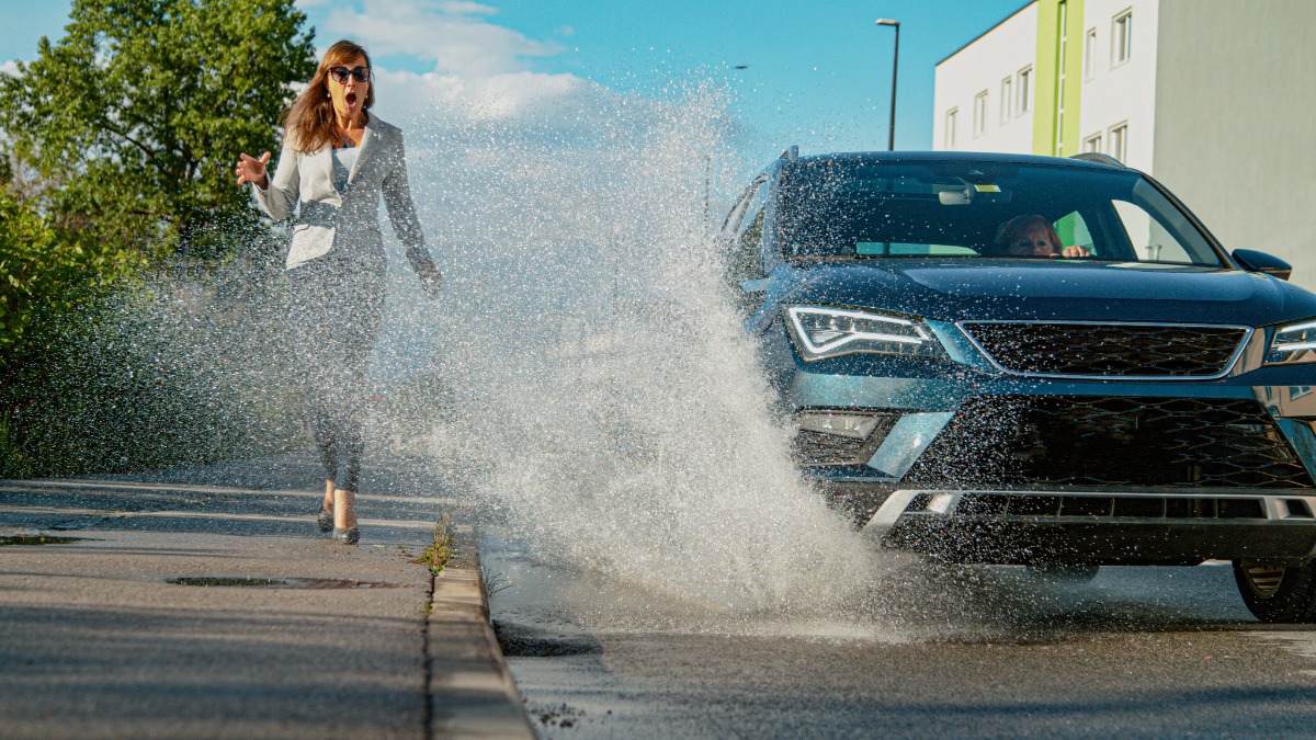 Auto fährt durch Pfütze und spritzt eine Fußgängerin nass