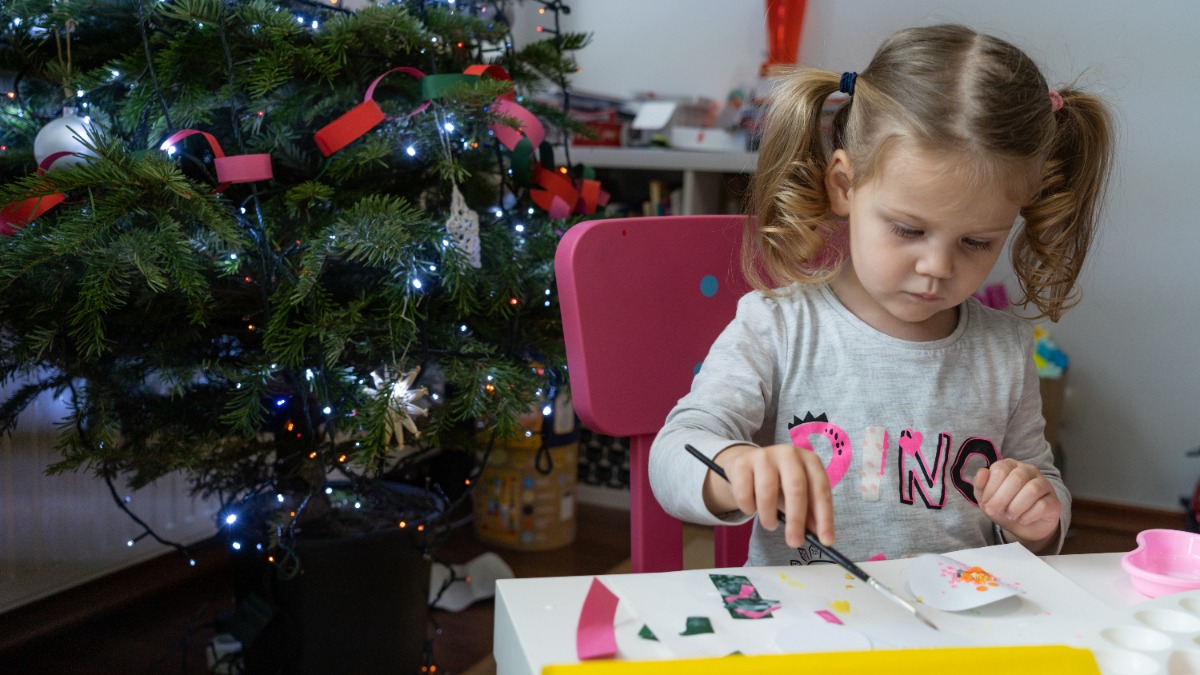 nützliche Geschenke Kinder: ein kleines Mädchen sitzt neben einem Weihnachtsbaum am Tisch und bastelt