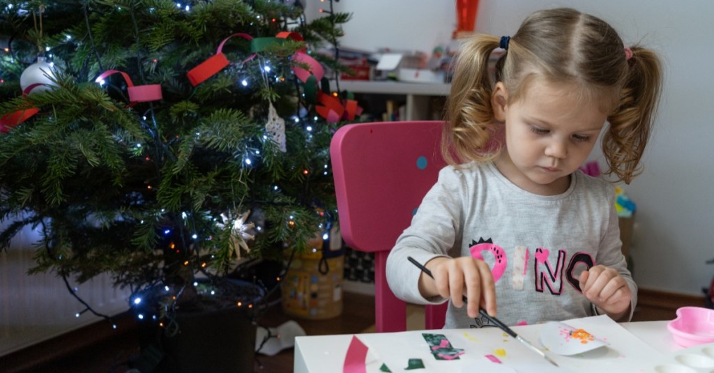 nützliche Geschenke Kinder: ein kleines Mädchen sitzt neben einem Weihnachtsbaum am Tisch und bastelt