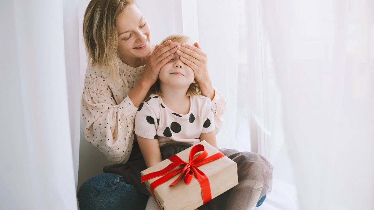 Alleinerziehend an Weihnachten: Mutter verdeckt die Augen ihrer Tochter, auf deren SchoÃŸ ein Geschenk liegt