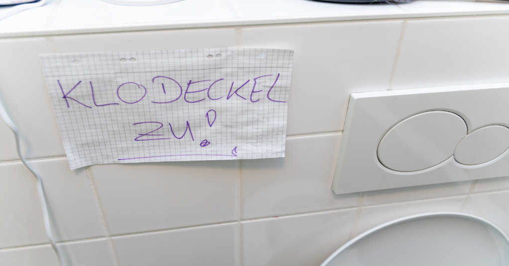 Ein Zettel mit der Aufschrift: Klodeckel zu klebt in einer Toilette.