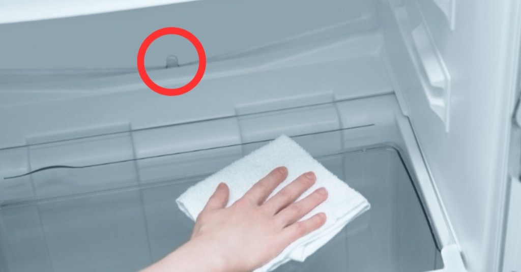 eine Hand wischt mit einem Lappen einen Kühlschrank sauber, der Wasserablauf ist mit einem roten Kreis markiert