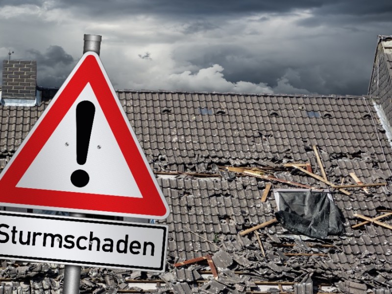 Ein Dach mit Sturmschäden im Hintergrund von einem Achtung-Verkehrszeichen mit der Aufschrift 
