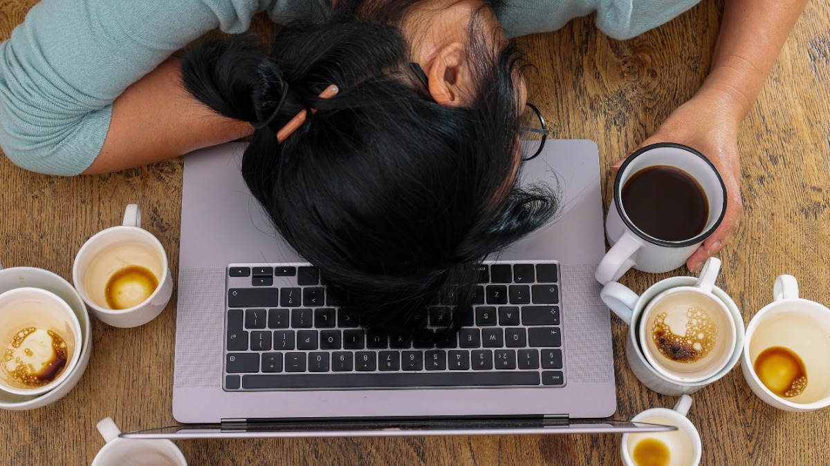 Eine Frau umgeben von leeren Tassen, die müde mit dem Kopf auf der Tastatur eines Laptops liegt.
